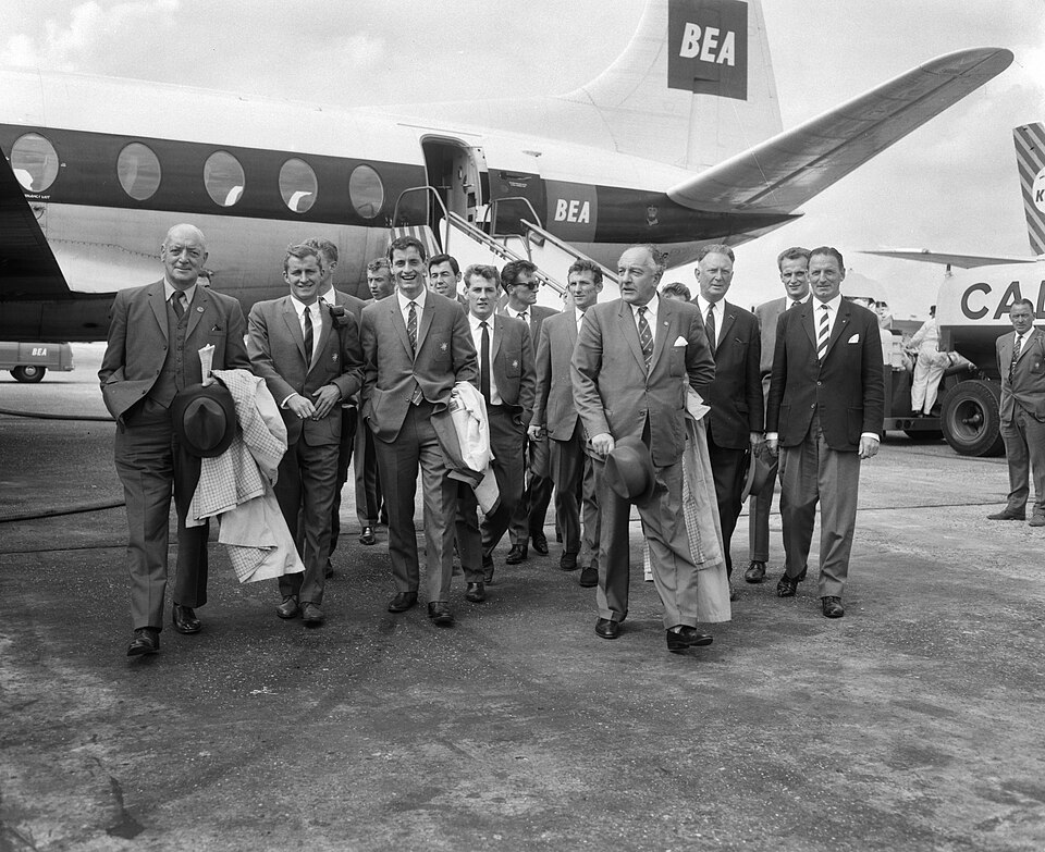 Footballers in suits walking across an airport apron beside an aircraft in 1963