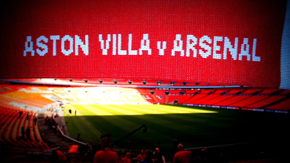 Wembley's big screen reading 'ASTON VILLA v ARSENAL' above the pitch, with empty red seats in the foreground
